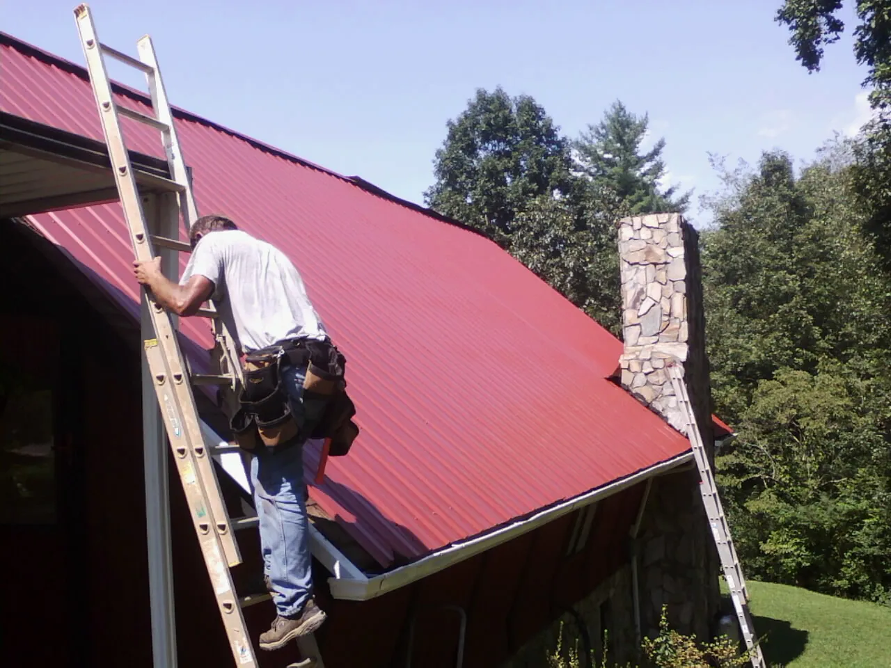 Roofer on a ladder beside a red metal roof and stone chimney — Soffit & Fascia Repair by Roofing Brothers in Lynbrook